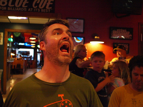 Front, chest up view of a person with their mouth wide open, in a dimly lit bar, with people watching them from behind